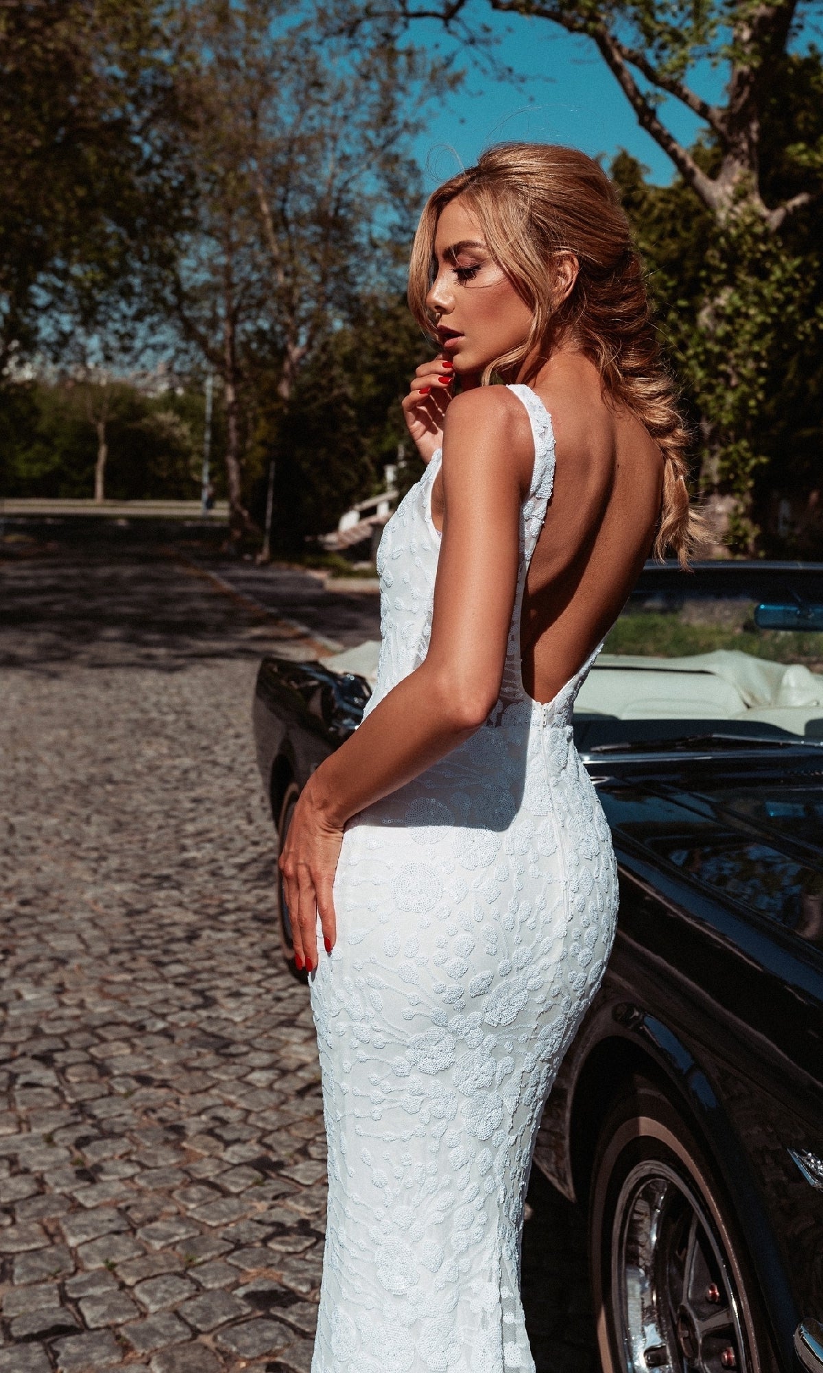 A woman in the Long White Mermaid Bridal Gown: Tina Holly BB011 stands beside a black convertible on a cobblestone road, with trees and sunlight in the background.